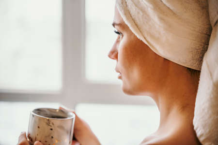 Young serene relaxed woman in spa bath towel drinking hot beverage tea coffee after taking shower bath at home. Beauty treatment, hydration concept.の写真素材