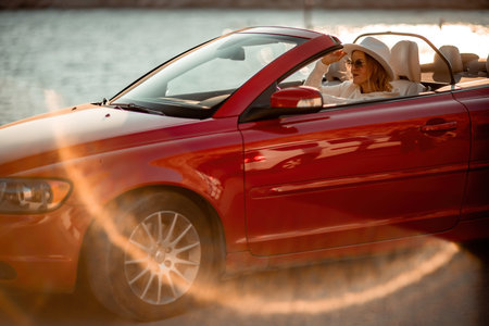 Outdoor summer portrait of stylish blonde woman driving red car convertible. Fashionable attractive woman with blond hair in a white hat in a red car. Sunny bright colors taken outdoors against the seaの写真素材
