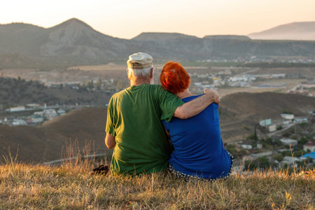 An elderly couple sits on a mountain with their backs with a beautiful view of the mountains and the sea in the distance.の写真素材