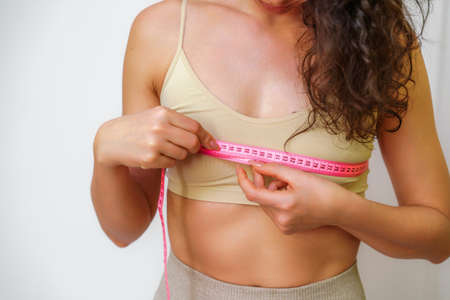 Cropped view of slim woman measuring breasts with tape measure at home, close up. Unrecognizable European woman checks the result of a weight loss diet or liposuction indoors. Healthy lifestyle.の写真素材