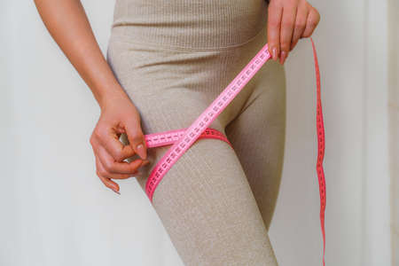 Cropped view of slim woman measuring her leg with tape measure at home, close-up. An unrecognizable European woman checks the result of a weight loss diet or liposuction indoors. Healthy lifestyle.の写真素材