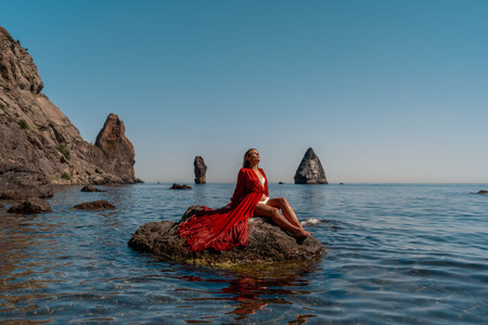 Beautiful sensual woman in a flying red dress and long hair, sitting on a rock above the beautiful...