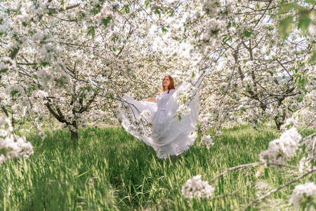 Fantasy woman in long white elegant fashion long dress walks in green spring blossom cherry garden. Happy cheerful girl princess bride. Skirt fabric flies flowing waving in wind motion.の写真素材
