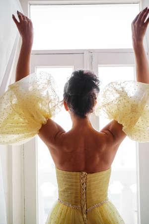 A womans silhouette in a golden luxurious dress against the background of a window holds a curtain with her hands. Elegant lady in a yellow long silk dress with bare back, back view.の写真素材