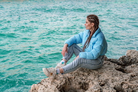 A woman in a blue jacket sits on a rock above a cliff above the sea, looking at the stormy ocean....
