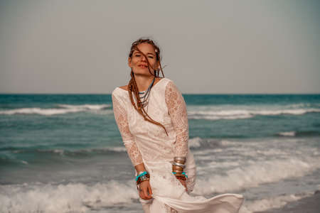 Model in boho style in a white long dress and silver jewelry on the beach. Her hair is braided, and there are many bracelets on her arms.の写真素材