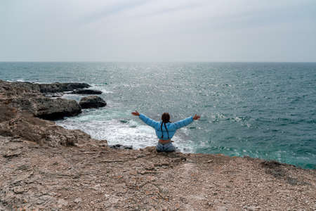 A woman in a blue jacket sits on a rock above a cliff above the sea, looking at the stormy ocean. Girl traveler rests, thinks, dreams, enjoys nature. Peace and calm landscape, windy weather.の写真素材