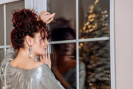 Woman portrait window bokeh christmas tree. A curly-haired middle-aged woman looks out of a dark...