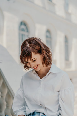 Business woman city street. A woman in a white shirt and a blue denim skirt sits on the parapet and holds a blue notebook in her hands against the background of the building. Summer city walks, travelの写真素材