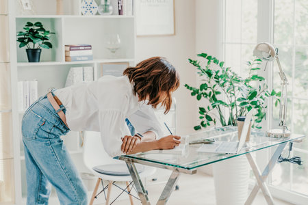 Business woman in bright office. A woman writes in a notebook while standing near the workplace desk. A business woman working at a desk in an office is dressed in jeans and a white shirt.の写真素材