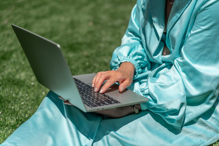 a young beautiful woman with blond curly hair in glasses and a blue dress sits on the grass in nature and uses a laptop,の写真素材