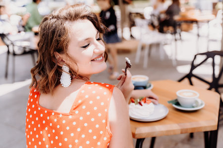 Charming woman in a restaurant, cafe on the street. She sits at the table and eats a cake with a fork. Dressed in a red sundress with white polka dots.の写真素材