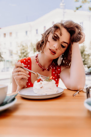Charming woman in a restaurant, cafe on the street. She sits at the table and eats a cake with a fork. Dressed in a red sundress with white polka dots.の写真素材