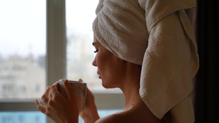 Middle aged woman looks good with bare shoulders in a white towel on her head holds a cup and drinks...
