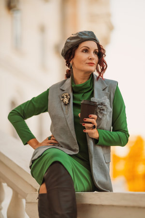 Outdoor fashion portrait of an elegant fashionable brunette model in a beret, green dress and a gray waistcoat posing at sunset in a european city in autumn.の写真素材