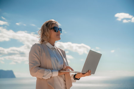 Freelance women sea. She is working on the computer. Good looking middle aged woman typing on a laptop keyboard outdoors with a beautiful sea view. The concept of remote work.の写真素材