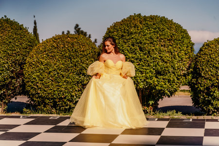 Woman yellow dress chess. A beautiful woman in a long puffy yellow dress poses on a chessboard in the park.の写真素材