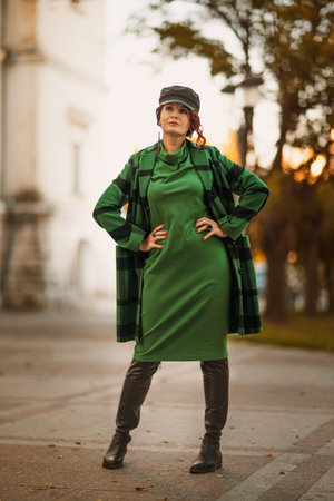 Outdoor fashion portrait of an elegant fashionable brunette woman, model in a stylish cap, green dress, posing at sunset in a European city in autumn.の写真素材