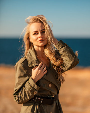 Portrait blonde sea cape. Calm young blonde in a khaki raincoat stands on the seashore