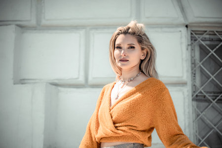 A middle-aged woman looks like a good blonde with curly beautiful hair and makeup on the background of the building. She is wearing a yellow sweater.の写真素材