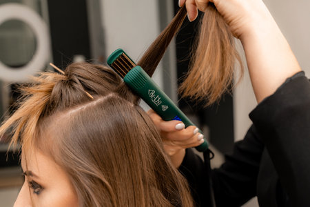 The hairdresser prepares a woman, makes a hairstyle with curls using a curling iron. Long light brown natural hair. Self care concept.の写真素材