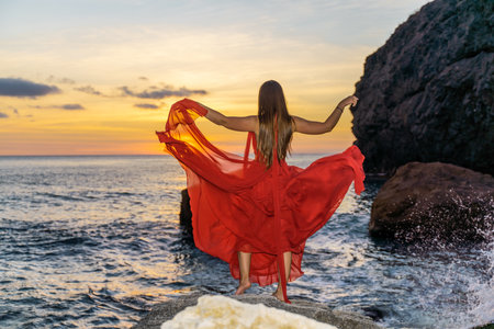 Woman in a red flying dress on the ocean or on the sea beach against the backdrop of the sunset sky.の写真素材