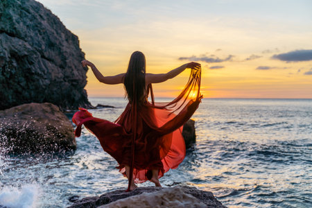 Woman in a red flying dress on the ocean or on the sea beach against the backdrop of the sunset sky.の写真素材