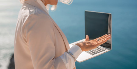 Freelance women sea working on the computer. Good looking middle aged woman typing on a laptop keyboard outdoors with a beautiful sea view. The concept of remote work.の写真素材