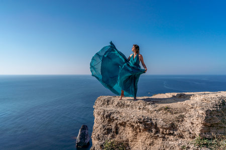 A girl with loose hair in a long mint dress descends the stairs between the yellow rocks overlooking the sea. A rock can be seen in the sea. Sunny path on the sea from the rising sunの写真素材
