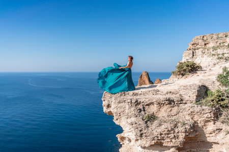 A girl with loose hair in a long mint dress descends the stairs between the yellow rocks overlooking the sea. A rock can be seen in the sea. Sunny path on the sea from the rising sunの写真素材