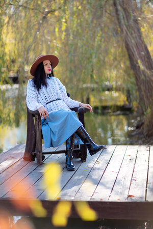 Autumn lake woman. She sits by a pond on a wooden pier in autumn and admires nature. The concept of tourism, weekends outside the city.の写真素材