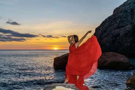 Woman in a red flying dress on the ocean or on the sea beach against the backdrop of the sunset sky.の写真素材