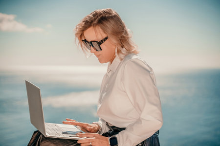 Business woman on nature in white shirt and black skirt. She works with a laptop in the open air with a beautiful view of the sea. The concept of remote work.の写真素材