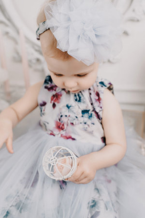 Baby girl elegant dress. A one-year-old girl in a puffy dress and a cute bow poses against the backdrop of a bright room with a dressing table and flowers.の写真素材