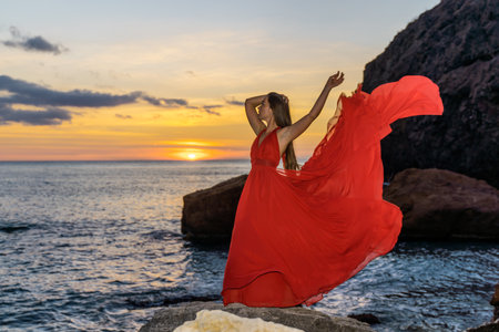 Woman in a red flying dress on the ocean or on the sea beach against the backdrop of the sunset sky.の写真素材