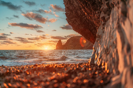 Sea sunset waves with foam. Waves run on pebbles and rock from volcanic basalt. Water breaks into splashes and white foam. Against the background of the cape and the sunset sky.の写真素材