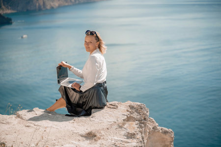 Business woman on nature in white shirt and black skirt. She works with a laptop in the open air with a beautiful view of the sea. The concept of remote work.の写真素材