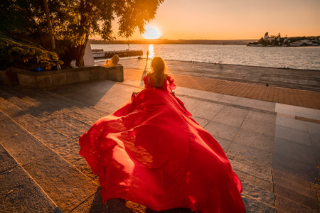 Sunrise red dress. A woman in a long red dress against the backdrop of sunrise, bright golden light of the suns rays. The concept of femininity, harmony.の写真素材