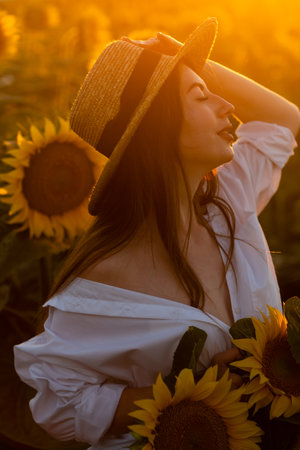 A girl in a hat on a beautiful field of sunflowers against the sky in the evening light of a summer sunset. Sunbeams through the flower field. Natural background.の写真素材