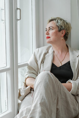 A middle-aged woman in a beige suit and black tank top sits mysteriously and looks out the window on...
