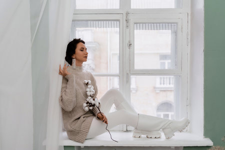 Free time for rest. Profile of a beautiful woman sitting on a white window sill at home, holding a cotton plant in her hands. She is wearing a sweater, leggings and white boots.の写真素材