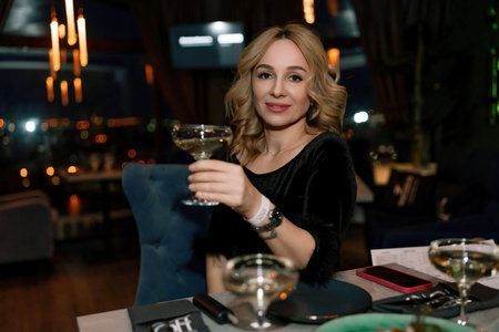 woman in a restaurant with champagne in her hand. She is sitting at a table against the backdrop of a dark restaurant hall with lamps.の写真素材