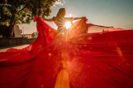 Sunrise red dress. A woman in a long red dress against the backdrop of sunrise, bright golden light of the suns rays. The concept of femininity, harmony.の写真素材