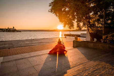 Sunrise red dress. A woman in a long red dress against the backdrop of sunrise, bright golden light of the suns rays. The concept of femininity, harmony.の写真素材