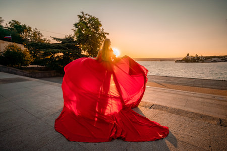 Sunrise red dress. A woman in a long red dress against the backdrop of sunrise, bright golden light of the suns rays. The concept of femininity, harmony.の写真素材