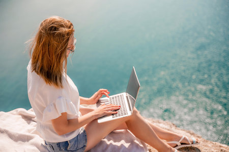 Freelance women sea working on the computer. Good looking middle aged woman typing on a laptop keyboard outdoors with a beautiful sea view. The concept of remote work.の写真素材