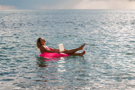 Woman works on laptop in sea. Freelancer, blond woman in sunglases floating on an inflatable big pink donut with a laptop in the sea at sunset. People summer vacation rest lifestyle concept.の写真素材