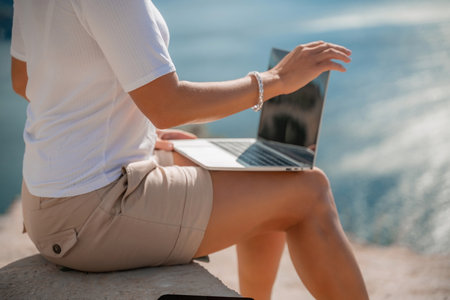 Freelance women sea working on the computer. Good looking middle aged woman typing on a laptop keyboard outdoors with a beautiful sea view. The concept of remote work.の写真素材