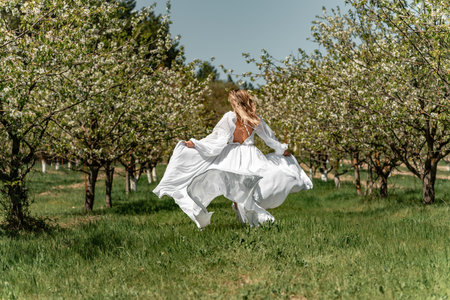 A woman in a white dress runs through a blossoming cherry orchard. The long dress flies to the sides, the bride runs rejoicing in life.の写真素材