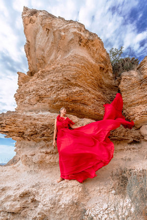 A woman in a red silk dress stands by the ocean, with mountains in the background, as her dress sways in the breeze.の写真素材